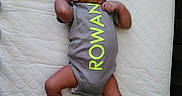Rowan is registered to the contest to win money with this photo: baby, infant, onesie, gray_clothing, neon_text, quilted_mattress, lying_down, newborn, curious_expression, small_hands, small_feet, child, person, indoor, soft_lighting, portrait, cute, sleeping_area, baby_clothes, resting