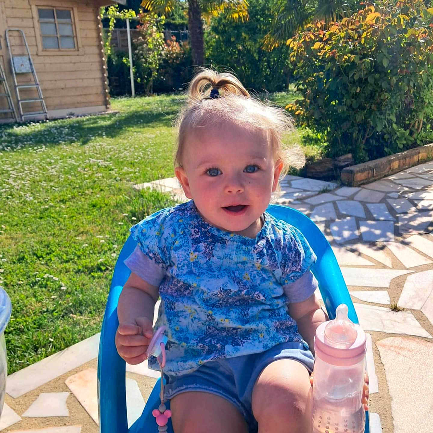 Luna a rejoint le concours — aidez-le/la à gagner de superbes lots ! baby_bottle, blue_chair, child, daytime, face, garden, grass, greenery, happy, outdoor, pacifier, pavement, playful, portrait, shorts, summer, sunlight, toddler, tshirt, wooden_shed