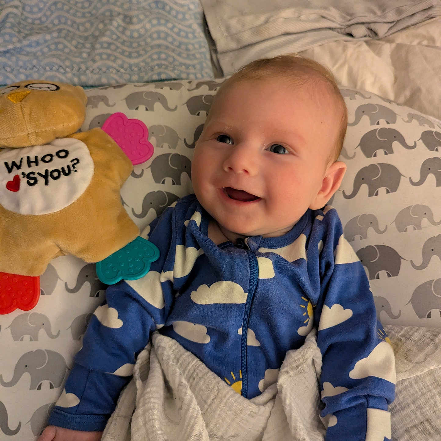 Holstin is registered to the contest to win money with this photo: baby, bed, blanket, blue_onesie, child, cloud_pattern, comfort, cozy, cushion, cute, elephant_pattern, face, happy, indoor, infant, person, plush_toy, sleepwear, smiling, soft_toy