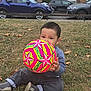 Daniyal is registered to the contest to win money with this photo: toddler, child, grass, soccer_ball, outdoor, parked_cars, autumn_leaves, sitting, playing, casual_clothing, short_hair, building, sidewalk, daytime, young_child, holding_ball, colorful, toy, nature, urban