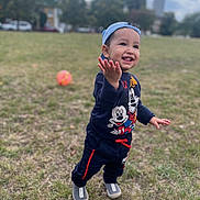 Daniyal is registered to the contest to win money with this photo: ball, blue_cap, cap, casual_clothing, child, daylight, gesture, grass, happy, mickey_mouse, outdoor, pants, park, person, playing, shoes, smiling, sweatshirt, toddler, young_child