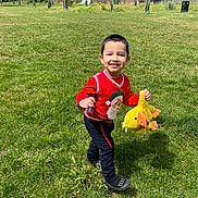 Daniyal joined the competition — help win amazing prizes! child, smiling, running, red_sweater, stuffed_toy, yellow_duck, grass, park, outdoor, daytime, happy, person, playing, toy, nature, casual_clothing, greenery, sunny, cute, young