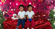 Donte is registered to the contest to win money with this photo: two_children, toddler, heart_balloons, valentines_day, red_bench, teddy_bear, white_tshirt, indoor, decorations, plants, carpet, smiling, curly_hair, casual_clothing, celebration, festive, cute, friendship, seated, colorful