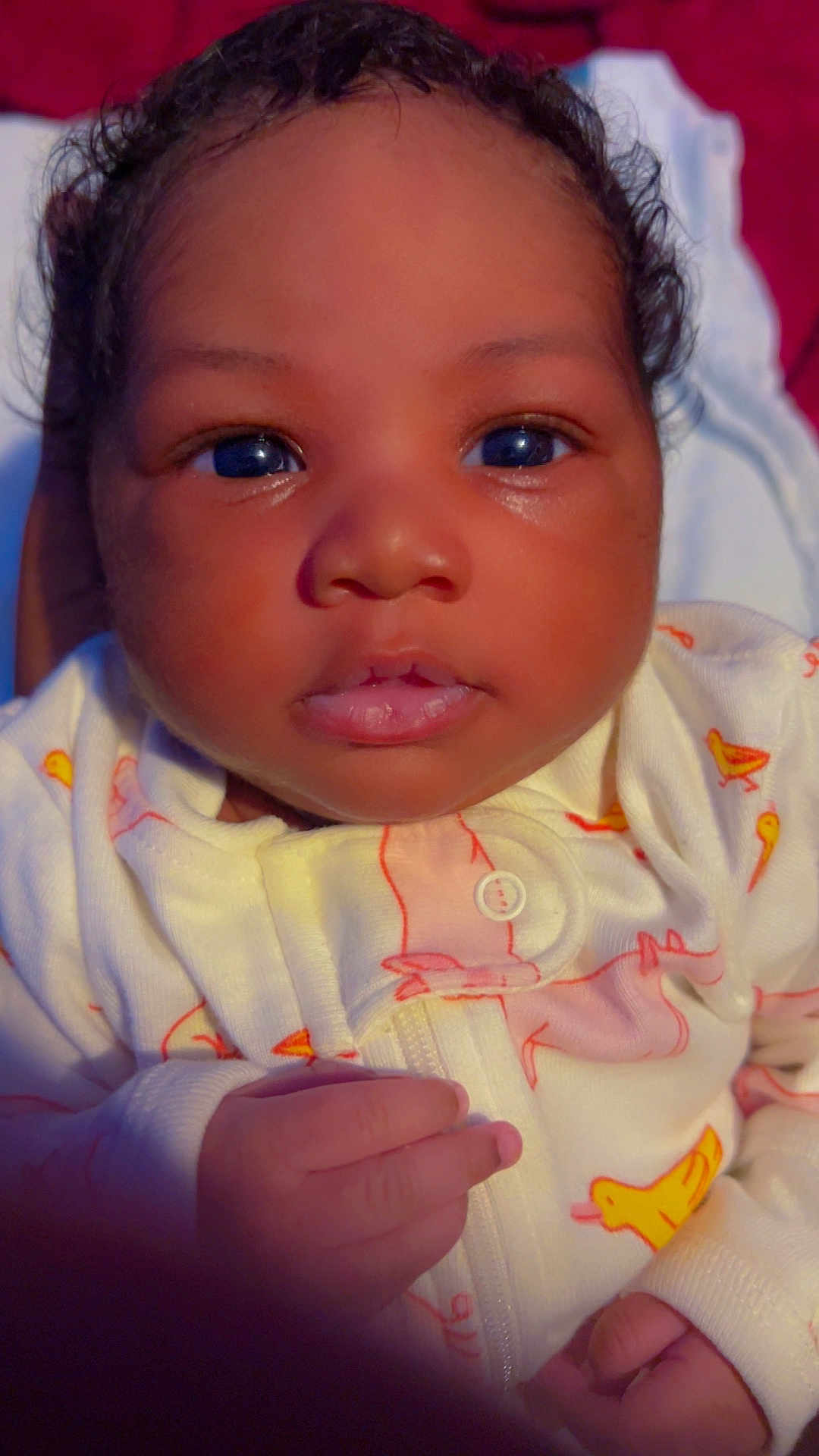 Riley is registered to the contest to win money with this photo: baby, infant, close_up, face, eyes, curly_hair, onesie, hand, cute, child, newborn, soft_skin, looking, portrait, indoors, cozy, sleepwear, yellow, pink, expression