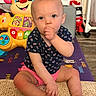 Zoey is registered to the contest to win money with this photo: baby, child, sitting, thumb_sucking, indoor, toy, colorful, blue_eyes, curious, pink_shorts, navy_shirt, patterned_clothing, playmat, carpet, floor, face, person, home, cute, infant