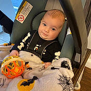 Louis is registered to the contest to win money with this photo: baby, infant, car_seat, blanket, toy, halloween, skeleton_outfit, smile, indoor, child, person, face, clothing, head, seatbelt, wood_floor, curious, cute, cozy, playful