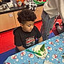 child, curly_hair, black_tshirt, table, tablecloth, snowman_pattern, paper_plate, green_treat, plastic_knife, classroom, holiday_decorations, floor_tiles, person, chair, cabinet, pencil_holder, cup, wrapping_paper, food, indoor