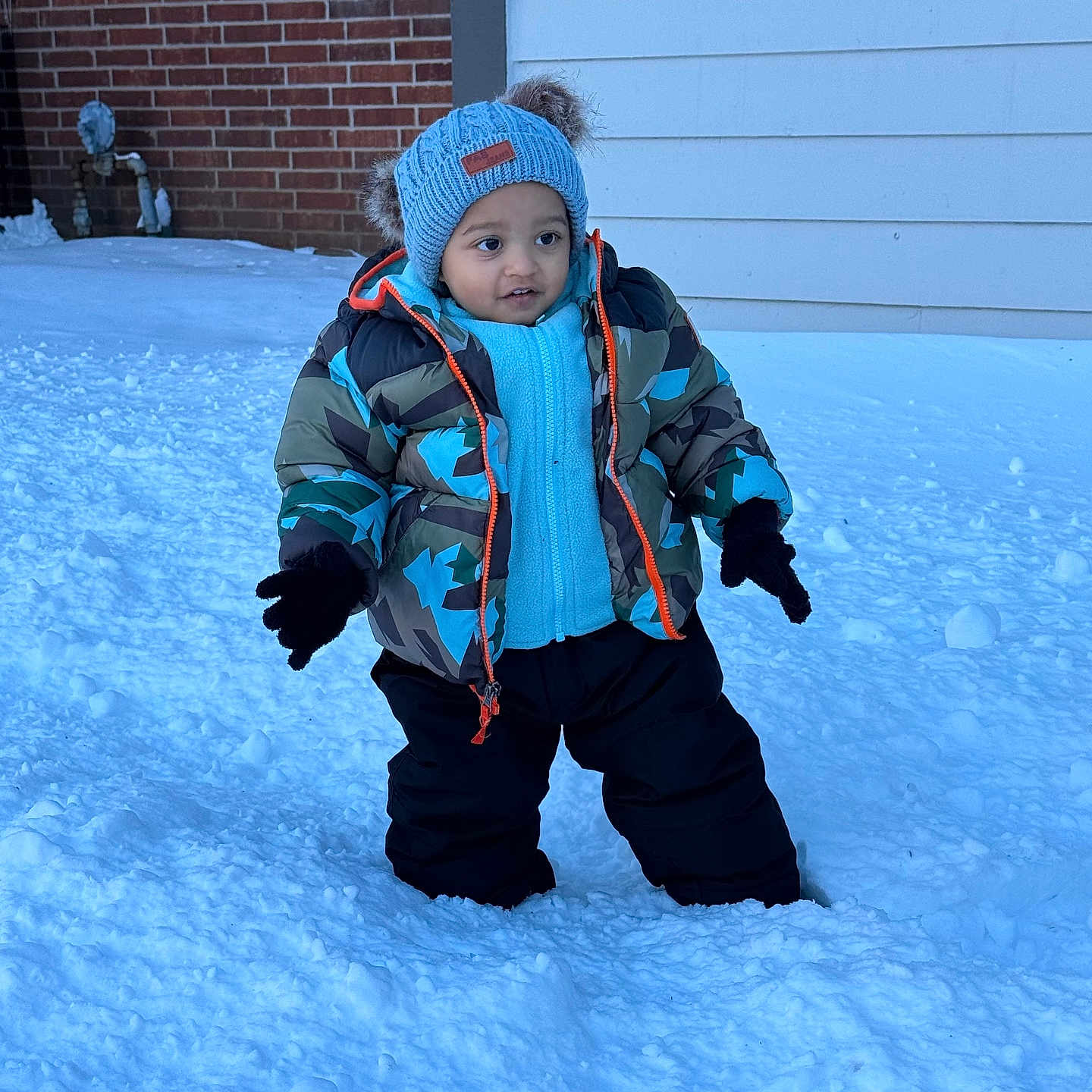 Rudransh is registered to the contest to win money with this photo: toddler, child, snow, winter_clothing, hat, jacket, gloves, outdoor, cold_weather, snow_pants, standing, house, brick_wall, window, curious, winter, footwear, blue_hat, camouflage_jacket, snowy_ground