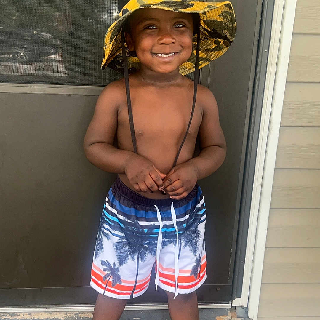 Zamari joined the competition — help win amazing prizes! bare_chest, black_shoes, boy, child, door, doormat, footwear, happy, hat, outdoor, palm_tree, person, porch, shorts, skin, smiling, standing, summer, toddler, yellow_hat