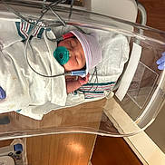 Kimberley joined the competition — help win amazing prizes! newborn, baby, hospital, bassinet, blanket, pacifier, hat, medical_equipment, gloves, sleeping, infant, person, bed, monitor, wooden_floor, hospital_room, swaddled, cute, healthcare, peaceful