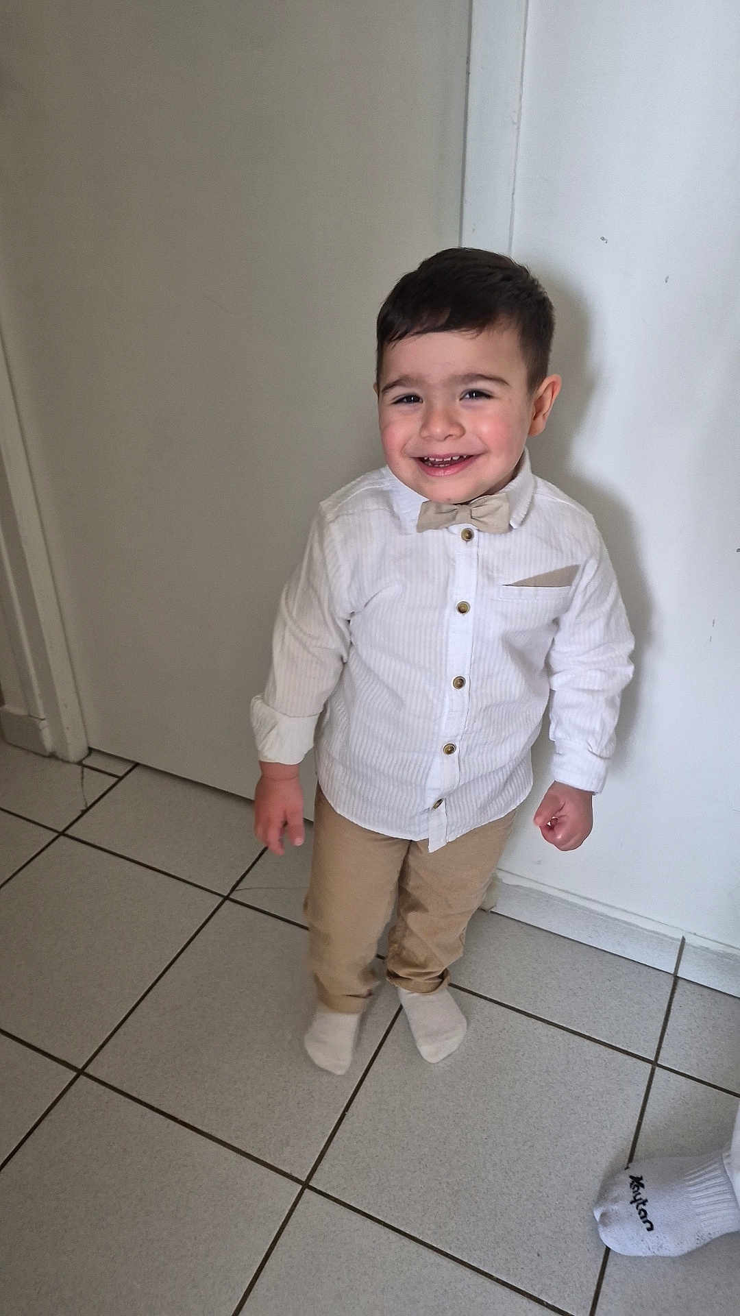 Adnan participe au concours pour gagner de l'argent avec cette photo : child, boy, toddler, smiling, bow_tie, white_shirt, beige_pants, socks, indoor, tiled_floor, doorway, wall, portrait, standing, happy, cute, short_hair, face, hands, feet