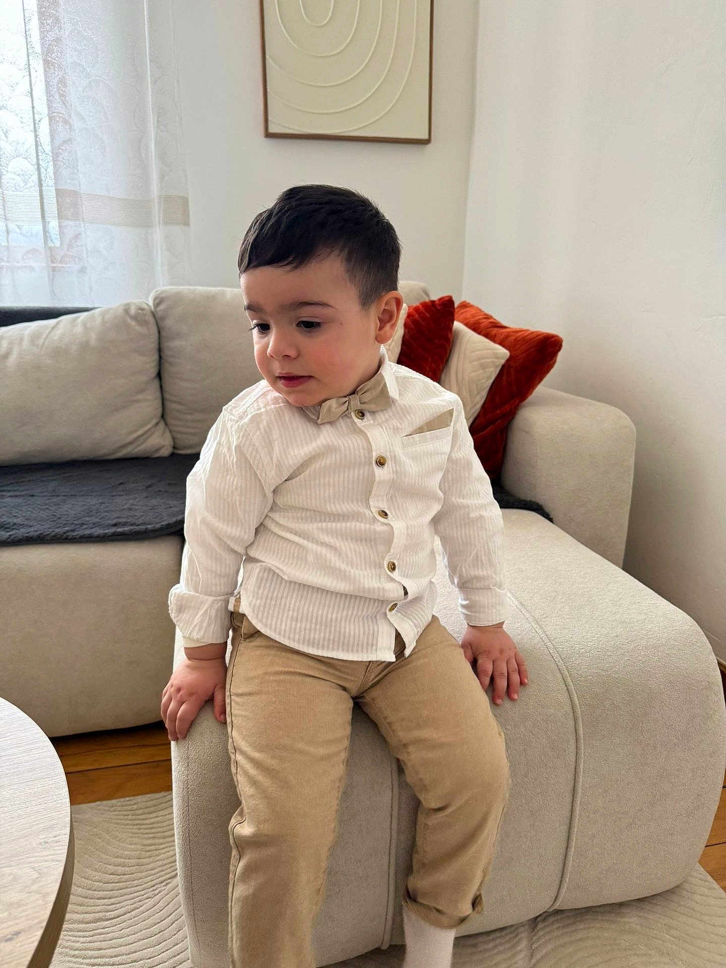 Adnan participe au concours pour gagner de l'argent avec cette photo : child, toddler, boy, bow_tie, white_shirt, beige_pants, sitting, sofa, ottoman, cushion, living_room, curtain, portrait, hands, short_hair, carpet, homedecor, cute, neutral_expression, indoor