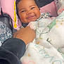 baby, smiling, curly_hair, blanket, car_seat, pink, black, hand, person, infant, happy, cozy, child, seatbelt, clothing, portrait, indoors, cute, comfort, toddler
