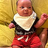baby, infant, child, bib, clothing, red_shorts, black_shirt, yellow_cushion, person, portrait, cute, indoors, sitting, expression, face, hand, foot, skin, text_on_clothing, relaxed