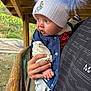 baby, child, hat, pom_pom, blue_eyes, jacket, hand, person, outdoor, wooden_structure, nature, curious, clothing, warm, infant, casual, holding, portrait, headwear, sleeve