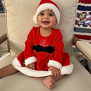 Emelia is registered to the contest to win money with this photo: baby, child, smiling, santa_hat, red_dress, christmas, holiday, indoor, chair, cushion, foot, hand, face, happy, festive, cute, person, portrait, cozy, home