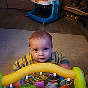 Jamarii is registered to the contest to win money with this photo: baby, child, toy, playtime, striped_clothing, indoor, carpet, walker, colorful, curious, floor, infant, seat, plastic_toy, rings, animal_toys, yellow, gray, person, young_child