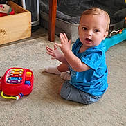 Jamarii joined the competition — help win amazing prizes! toddler, child, toy, carpet, blue_shirt, clapping, indoor, floor, play, person, furniture, wooden_box, heater, dog_bed, sock, shorts, curtains, colorful_toy, expression, home