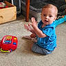 blue_shirt, carpet, child, clapping, colorful_toy, curtains, dog_bed, expression, floor, furniture, heater, home, indoor, person, play, shorts, sock, toddler, toy, wooden_box
