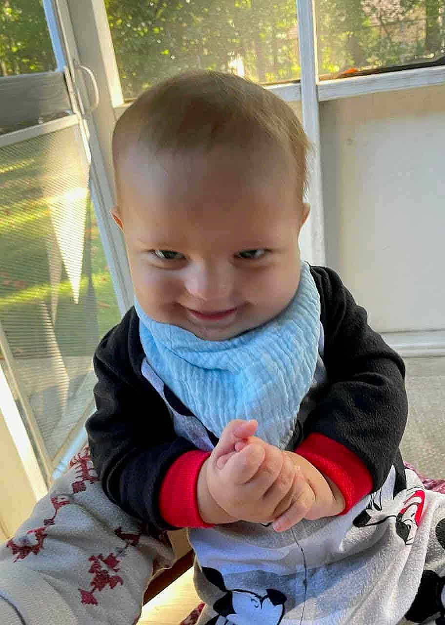 Lamarri is registered to the contest to win money with this photo: baby, child, indoor, window, sunlight, pajamas, blue_bib, hands, clothing, face, smile, expression, person, young_child, fingers, seat, background, room, light, casual