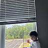 child, toddler, window, window_sill, indoor, blinds, shorts, tshirt, barefoot, hair, face, hand, legs, natural_light, curious, seated, daylight, greenery, parking_lot, casual_clothing