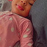 child, baby, pink_clothing, polka_dots, pillow, lying_down, face, portrait, resting, indoor, cozy, young_child, soft_expression, bed, blanket, head, close_up, casual, sleepwear, relaxed