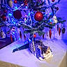 cat, kitten, sleeping, christmas_tree, christmas, ornaments, baubles, lights, tinsel, holiday, festive, indoor, table, tablecloth, decoration, blue_lighting, red_ornaments, cozy, relaxed, pet