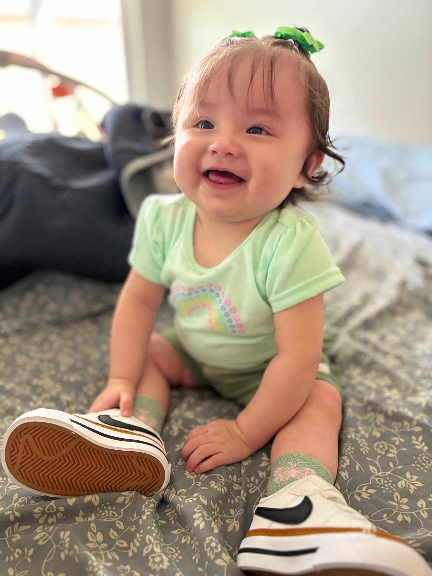 Kaia is registered to the contest to win money with this photo: baby, smiling, indoor, child, sitting, green_clothing, white_sneakers, patterned_bedspread, happy, cute, portrait, young_child, footwear, socks, tongue_out, natural_light, short_sleeves, hair_tie, infant, closeup