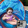 toddler, child, swimsuit, inflatable_pool, pool_float, canopy, smartphone, water, backyard, summer, outdoor, trees, fence, baby, sitting, concentration, play, fun, sunny, recreation