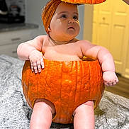Amina is registered to the contest to win money with this photo: baby, pumpkin, baby_in_pumpkin, kitchen, orange, cute, child, infant, costume, holiday, autumn, portrait, indoor, table, marble_countertop, headwear, curious, chubby, skin, hand