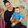baby, blue_shirt, casual_clothing, child, colorful_background, family, flower_pattern, hair_bun, happy, holding, indoor, mustard_pants, parent_and_child, person, portrait, seated, smile, sneakers, studio_photo, woman