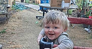 Aladar is registered to the contest to win money with this photo: box, chair, child, daytime, dirt_ground, fence, garden_tools, happy, outdoor, person, plants, playpen, potted_plants, smiling, sunlight, table, toddler, toys, umbrella, yard