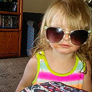 Aala is registered to the contest to win money with this photo: child, sunglasses, tank_top, blond_hair, living_room, carpet, cushion, patterned, tv, furniture, photo_frames, cabinet, person, indoor, casual, messy_hair, cute, colorful_clothing, home, portrait