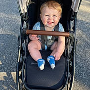 Leo is registered to the contest to win money with this photo: baby, child, stroller, smiling, blue_eyes, socks, outdoor, sunlight, pavement, happiness, infant, casual_clothing, seatbelt, sunshine, cute, portrait, happy, person, baby_clothes, daylight