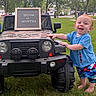 toddler, child, toy_jeep, grass, outdoor, park, trees, sign, blue_shirt, colorful_shorts, barefoot, smiling, red_hair, daylight, suburban, milestone, happy, person, play, nature