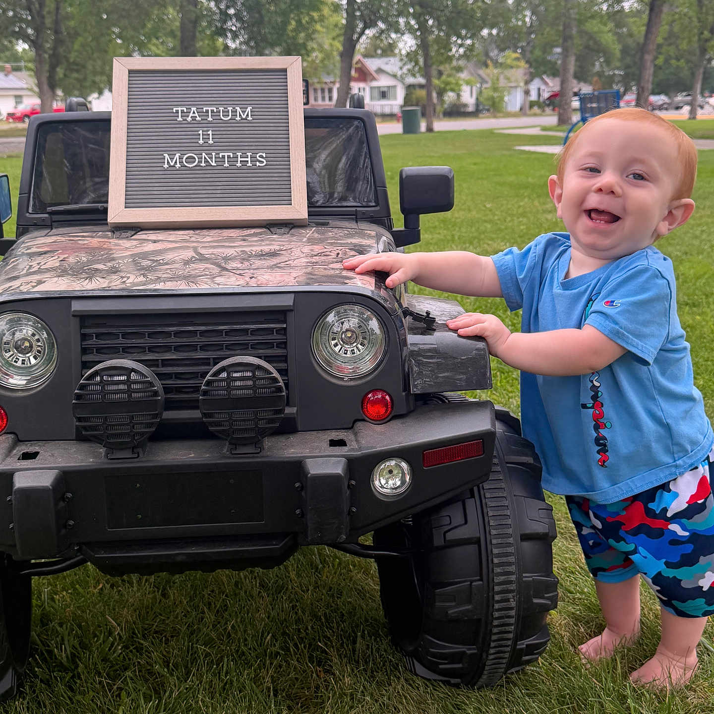Tatum is registered to the contest to win money with this photo: toddler, child, toy_jeep, grass, outdoor, park, trees, sign, blue_shirt, colorful_shorts, barefoot, smiling, red_hair, daylight, suburban, milestone, happy, person, play, nature
