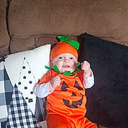 Asher is registered to the contest to win money with this photo: baby, infant, pumpkin_costume, halloween, couch, pillow, orange_outfit, smile, happy, costume, hat, green_leaf, jack_o_lantern, plush, sitting, indoor, portrait, eyes, fabric, cute