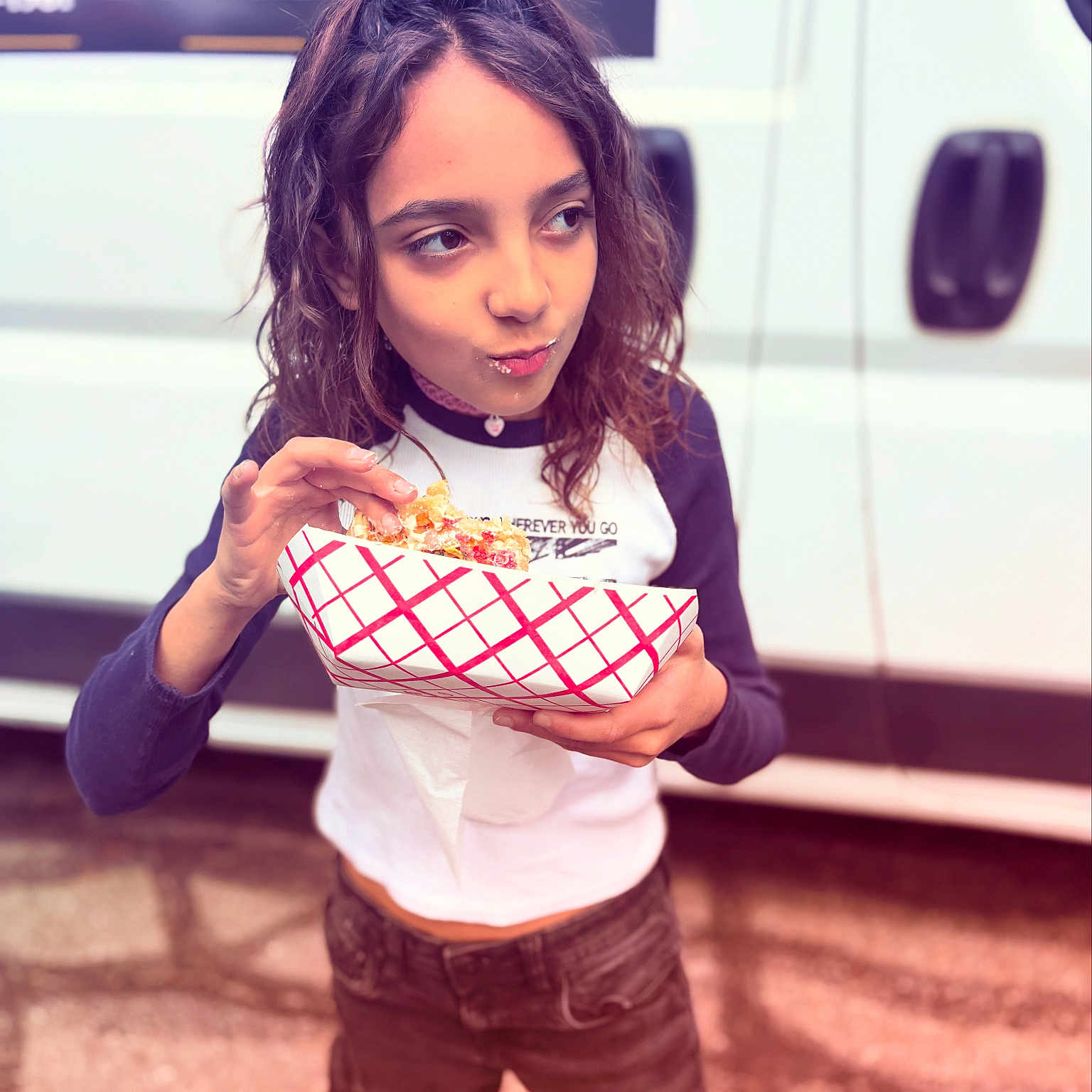 Penelope is registered to the contest to win money with this photo: background, casual_clothing, child, crumbs, curly_hair, daylight, eating, food, long_sleeve_shirt, menu_board, outdoor, pants, person, red_checkered, snack, standing, street, tray, van, white