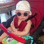 baby, child, sunglasses, hat, stroller, red, toy, book, floor, wooden_floor, table, cushions, indoor, person, clothing, foot, hand, seatbelt, playful, cute