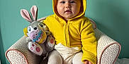 Leïya participe au concours pour gagner de l'argent avec cette photo : armchair, baby, bunny, child, colorful, costume, cozy, cute, easter_egg, footwear, indoor, orange_slippers, playful, plush_toy, portrait, seated, smiling, soft_toys, white_tights, yellow_hoodie