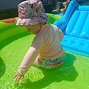 Adèle a rejoint le concours — aidez-le/la à gagner de superbes lots ! toddler, child, swimwear, bucket_hat, inflatable_pool, water, outdoor, sunlight, summer, play, blue_inflatable, dinosaur, person, table, chair, adult, backyard, grass, footwear, curious