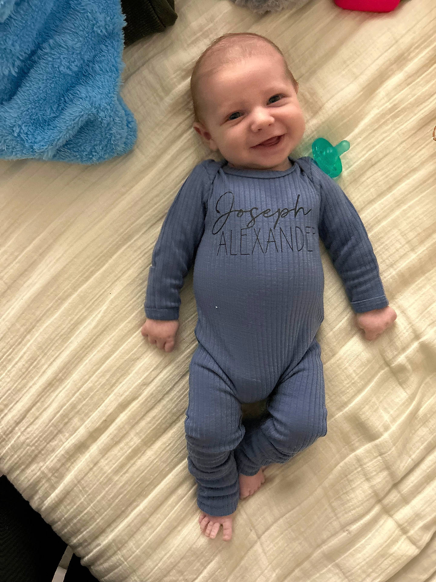 Joseph joined the competition — help win amazing prizes! baby, bed, blanket, blue_onesie, child, clothing, cute, face, hand, happy, head, indoor, infant, lying_down, newborn, pacifier, person, skin, smiling, towel