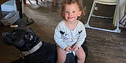 Zuri is registered to the contest to win money with this photo: toddler, child, dog, black_dog, curly_hair, pink_crocs, indoor, wooden_floor, high_chair, fan, dining_table, chair, smile, happy, pet, living_room, baby_clothes, floor, person, household