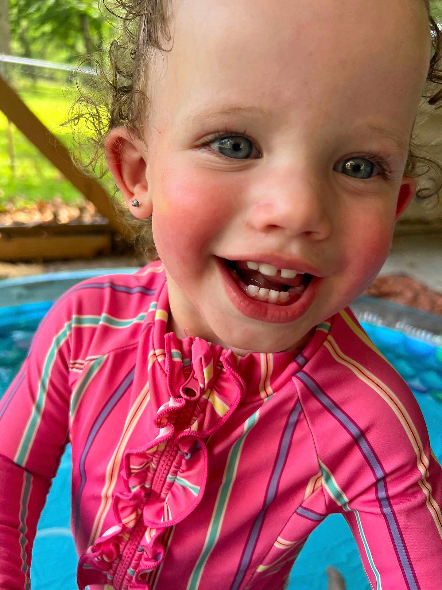 Zuri is registered to the contest to win money with this photo: child, toddler, face, smile, curly_hair, blue_eyes, pink_swimsuit, striped_clothing, ruffles, outdoor, kiddie_pool, water, happy, earring, closeup, summer, playful, person, skin, cheeks