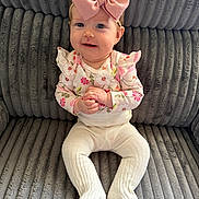 Kaija joined the competition — help win amazing prizes! baby, child, infant, pink_bow, floral_top, white_tights, smiling, sitting, couch, plush_fabric, indoor, cute, portrait, hands_clasped, headband, young_child, happy, person, cozy, texture