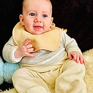 Roméo participe au concours pour gagner de l'argent avec cette photo : baby, infant, child, bib, sitting, smiling, blue_eyes, plush_toy, fleece_blanket, beige_clothing, onesie, portrait, indoor, close_up, adorable, chubby_cheeks, hands, feet, soft_texture, black_background