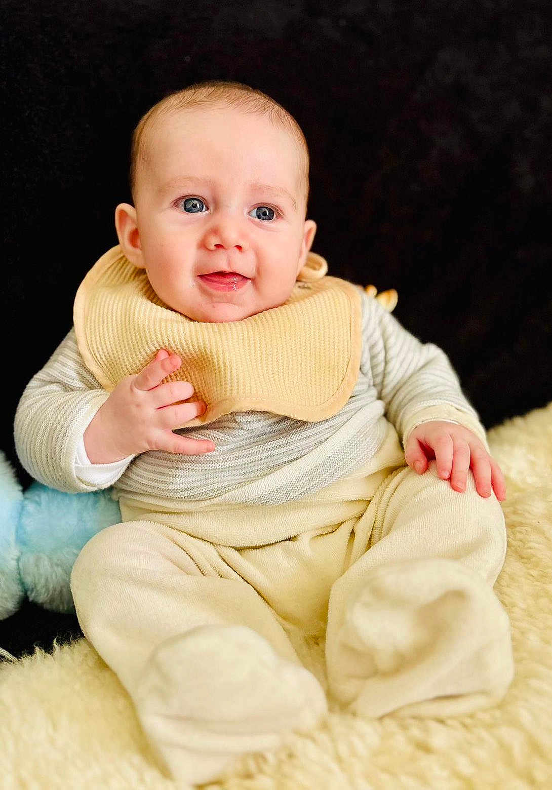 Roméo participe au concours pour gagner de l'argent avec cette photo : baby, infant, child, bib, sitting, smiling, blue_eyes, plush_toy, fleece_blanket, beige_clothing, onesie, portrait, indoor, close_up, adorable, chubby_cheeks, hands, feet, soft_texture, black_background