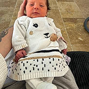 Kataleya a rejoint le concours — aidez-le/la à gagner de superbes lots ! newborn, baby, infant, child, sweater, white_clothing, animal_face, hand, tattoo, indoor, floor, tile_floor, seated, cozy, resting, cute, wearing, clothing, person, portrait