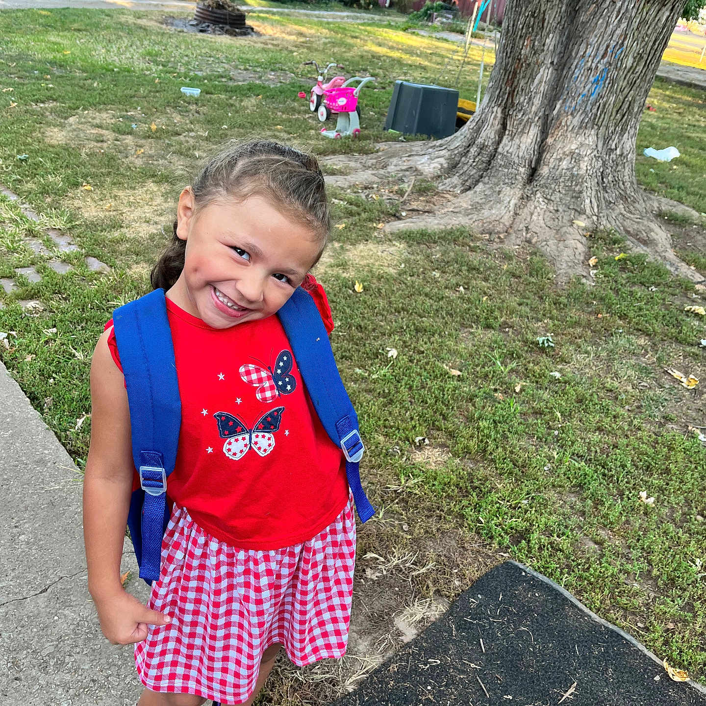 Kinzley is registered to the contest to win money with this photo: child, girl, smile, backpack, red_dress, braided_hair, outdoor, sidewalk, tree, grass, playground, tricycle, sunlight, happy, casual_clothing, nature, summer, play, portrait, cute