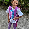 child, girl, backpack, purple, leggings, tshirt, cartoon, outdoor, gravel, smile, standing, nature, greenery, casual_clothing, footwear, confident, young, playful, portrait, person